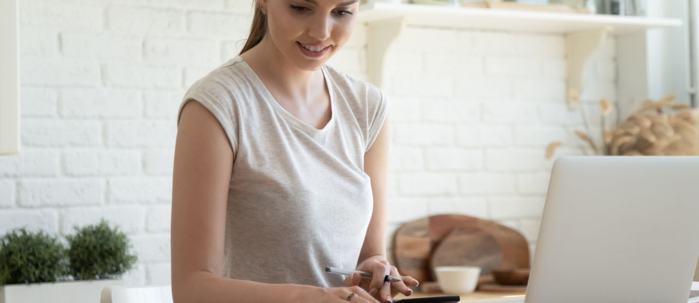 Smiling,Young,Woman,Checking,Finances,,Using,Laptop,,Sitting,At,Table