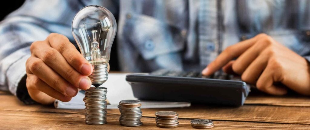 seseorang sedang menghitung sesuatu di kalkulator dan memegang bola lampu di atas tumpukan koin seseorang sedang menghitung sesuatu di kalkulator dan memegang bola lampu di atas tumpukan koin