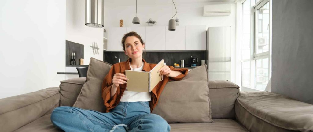 wanita duduk di sofa sambil membuka buku di dalam ruangan ber-ac wanita duduk di sofa sambil membuka buku di dalam ruangan ber-ac