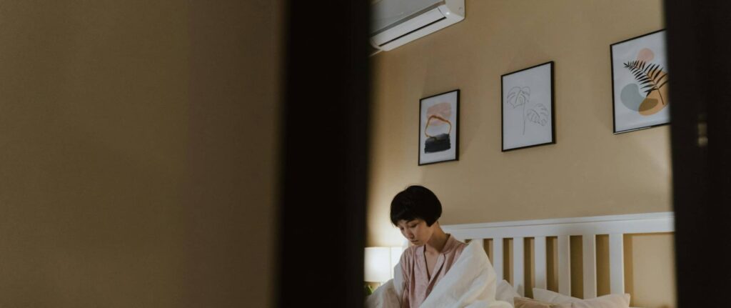 seorang wanita menggunakan selimut duduk di atas kasur dan ada unit AC di atas ranjang