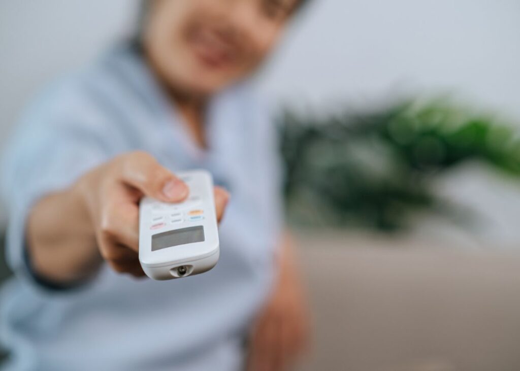 Seseorang mengarahkan remote AC berwarna putih ke depan dengan latar belakang yang buram