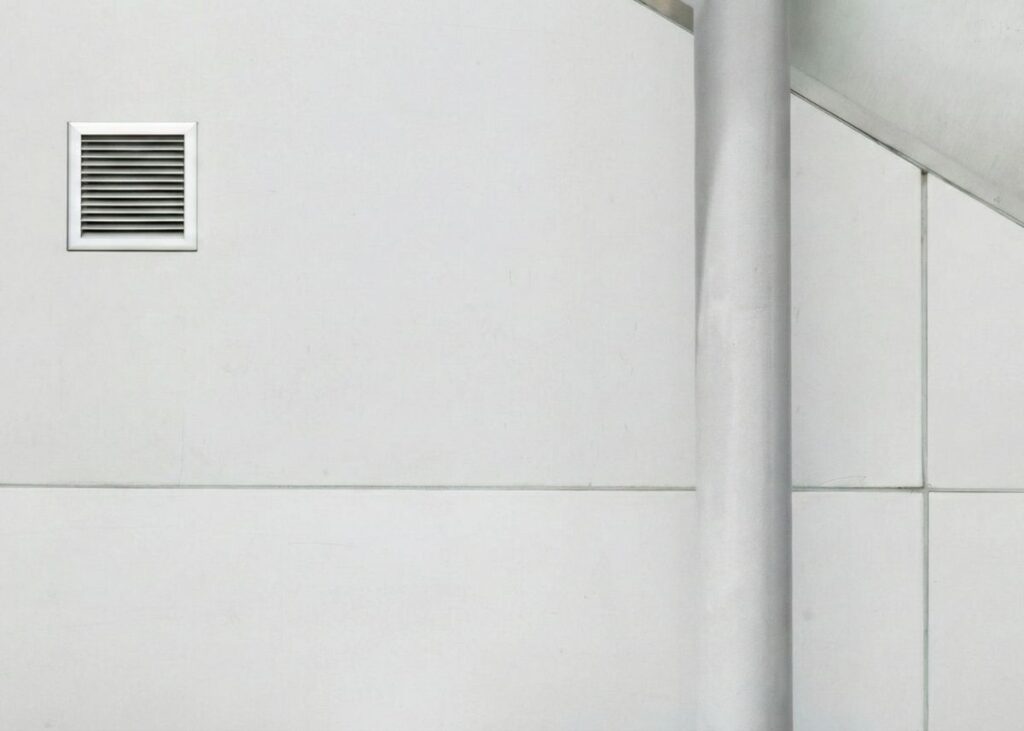 Dinding putih minimalis dengan lubang ventilasi kecil di kiri dan tiang vertikal di kanan